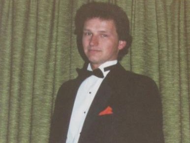 Man in black tuxedo and bow tie, orange pocket square, standing before green curtains