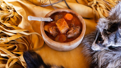 Wooden bowl of meat stew with carrots, antler-handled spoon on tanned leather and fur pelt