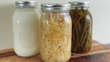 Three mason jars on wooden board: sauerkraut in center, white ferment on left, pickled green beans on right
