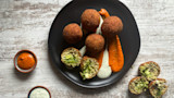 Five fried arancini on black plate with orange and white sauce streaks; two halved show green filling