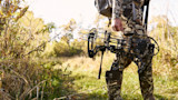 Hunter holding a compound bow with multiple arrows standing in grassy brush