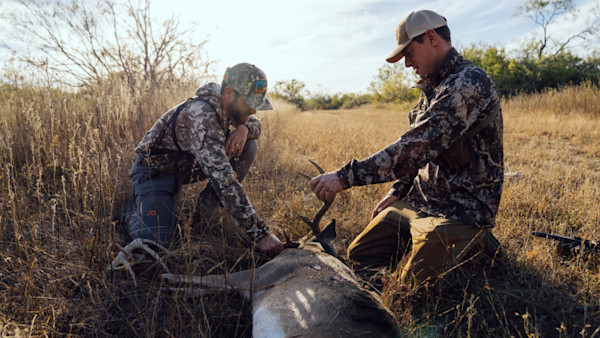 Two hunters kneel beside a downed whitetail deer in tall grass, one holding an antler; rifle on ground.