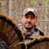 Man holding turkey tail fan, wearing camo jacket and cap in bare-branch woods