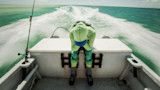 Seasick person hunched over stern of speeding boat, wearing green hoodie and blue cap