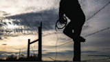 Hunter silhouette climbing barbed-wire fence at sunset holding antlers