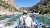 Two anglers in a raft on a fast canyon river, one facing away and one casting a fly rod