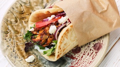 Mushroom gyro in pita with tomato, red onion, feta and greens, wrapped in brown paper on ceramic plate