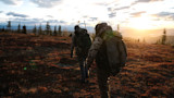 Two hunters walking away across tundra at sunset with backpacks and a rifle