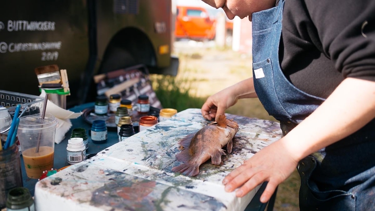 Person in denim apron arranging fish on paint‑splattered table with paint jars; text 'CHRISTIANSHINE 2019' on van