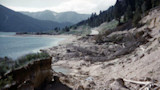 Eroded lake shoreline with collapsed banks and fallen trees; winding road and distant mountains.