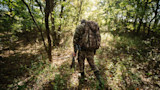 Hunter walking away in sunlit forest, wearing camo, carrying large pack and compound bow