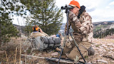 Two hunters on a ridge—one kneeling at binoculars on tripod, other reclining with backpack