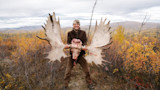 Hunter smiling, holding moose skull and large antlers in autumn tundra
