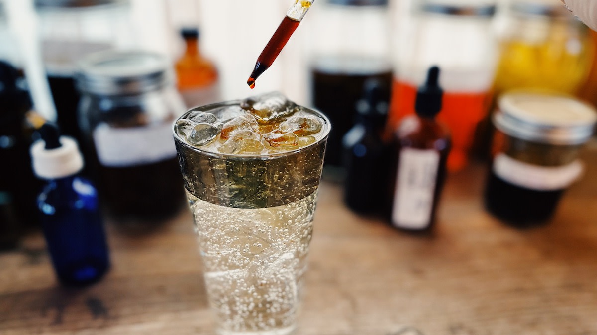 Glass of sparkling drink with ice and pipette dropping a drop of bitters, blurred bottles behind