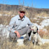 Man kneeling beside white-and-black hunting dog in grassy field with snow patches