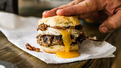 Biscuit sandwich with specklebelly boudin patty, fried egg oozing yolk, caramelized onions, hand pressing top