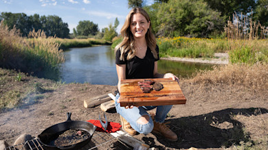 Woman kneeling by river holding cutting board with seared venison heart and cast-iron pan nearby