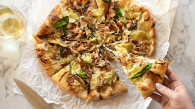 Pizza with shredded meat, artichoke hearts and greens; slice lifted by hand, glass of white wine