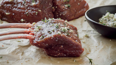 Raw rack of meat with exposed bones, sprinkled with herbs and coarse salt, bowl of herb mix