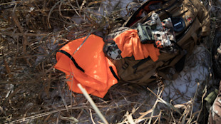 Blaze-orange hunting vest and camo backpack with binoculars and Canon lens cap lying in dry grass
