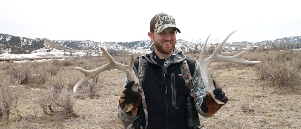 Hunter holding two shed antlers, wearing YETI cap, dry grass and snow-capped hills behind