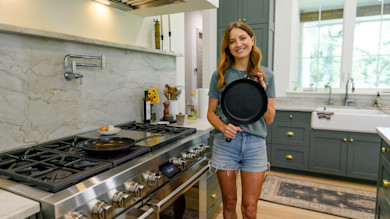 Smiling woman holding cast-iron skillet beside stovetop in bright kitchen