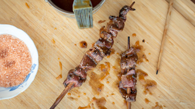 Guava-glazed venison heart skewers on wooden board with sauce brush and bowl of pink salt