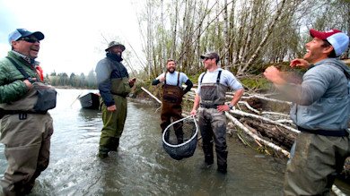 Five men in waders stand in a river, one holding a large fishing net with a boat behind