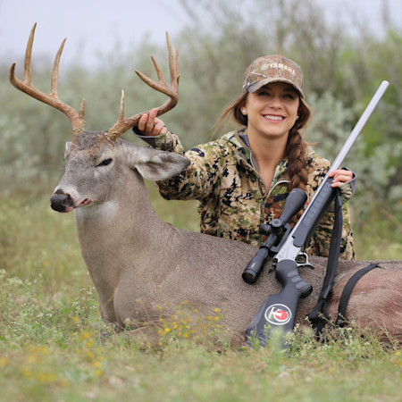 Smiling woman in camouflage holding buck antlers with rifle across deer; cap reads YAMAHA