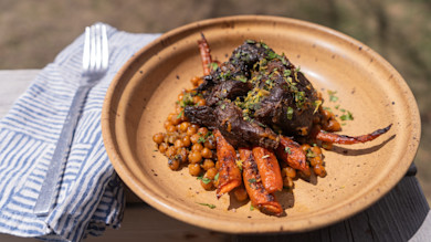 Braised meat over roasted carrots and chickpeas, herb garnish in tan bowl on wooden table