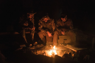 Three hunters sitting around a campfire grilling over a grate at night