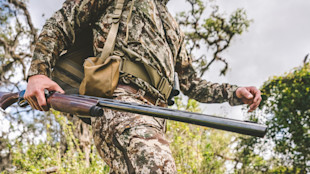 Over-under shotgun carried by hunter in camouflage walking near trees