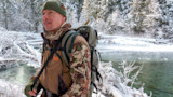 Hunter in First Lite camo jacket and green beanie beside a snowy creek
