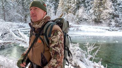 Hunter in First Lite camo jacket and green beanie beside a snowy creek