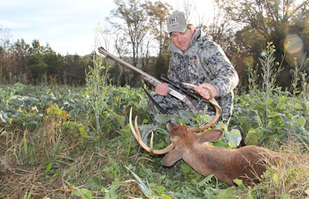 Hunter kneeling with rifle holding buck antlers beside harvested deer in a field