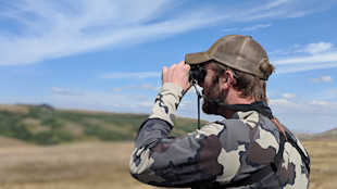 Hunter in camouflage jacket and cap looking through binoculars over grassland