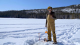 Hunter on frozen lake by an ice hole and auger, wearing brown bib overalls and fur hat