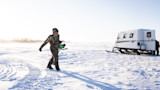 Hunter in camouflage bibs carrying green ice auger across sunlit frozen lake toward white snowcat