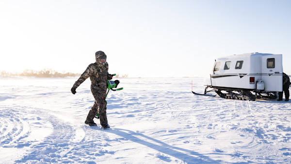 Hunter in camouflage bibs carrying green ice auger across sunlit frozen lake toward white snowcat