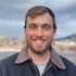 Smiling man with short brown hair and trimmed beard, wearing jacket, blurred lakeside background