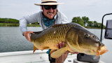 Smiling man on boat holding large carp toward camera on a lake