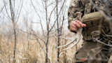 Hunter kneeling in dry grass holding a deer shed antler, camouflage gear and Vortex pouch