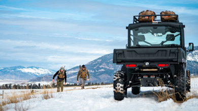 Two hunters walking across snowy field toward parked UTV with mountains behind