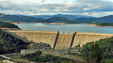Concrete dam with reservoir, forested hills and distant mountains under cloudy sky