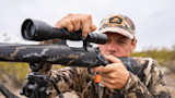 Hunter adjusting a rifle scope on a bipod, wearing camouflage and a cap with First Lite logo