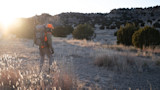 Hunter in orange vest and cap carrying pack and rifle walking toward sunrise across grassy hills