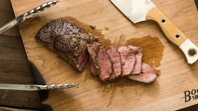 Smoked venison backstrap sliced on wooden cutting board with knife and metal tongs