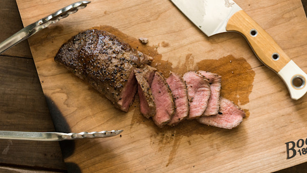 Smoked venison backstrap sliced on wooden cutting board with knife and metal tongs