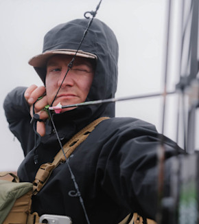 Hunter drawing a compound bow, wearing hooded jacket and cap, pink-fletched arrow nocked