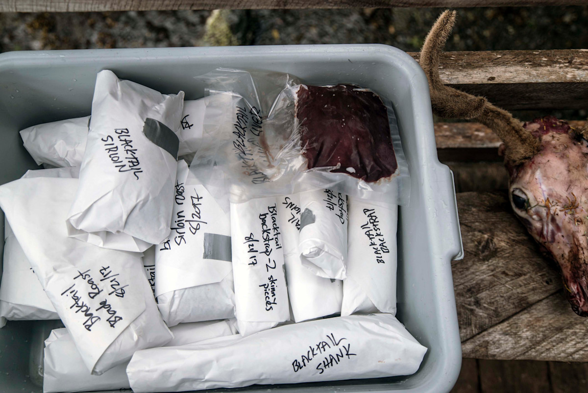Plastic tub of wrapped game meat labeled 'BLACKTAIL SIRLOIN' and 'BLACKTAIL SHANK', deer head nearby
